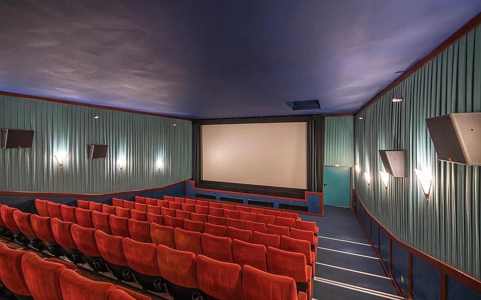 Empty red-seat cinema auditorium with a wide projection screen and wall speakers.